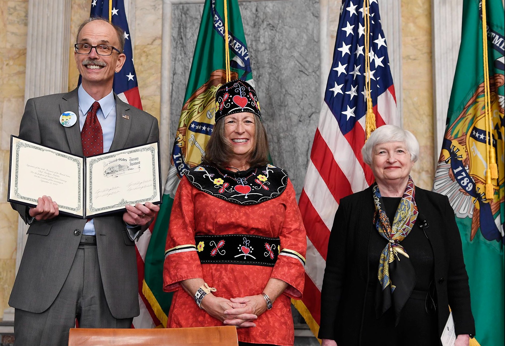 Director Jen Olijar, Treasurer Lynn Malerba, and Secretary Janet Yellen standing together in front of American and Treasury Flags. Len Olijar is holding a document showing Lynn Malerba's signature.
